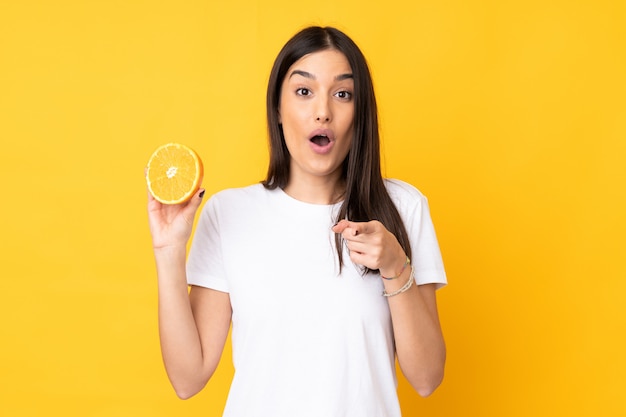 Mujer caucásica joven que sostiene una naranja en la pared amarilla sorprendida y que señala