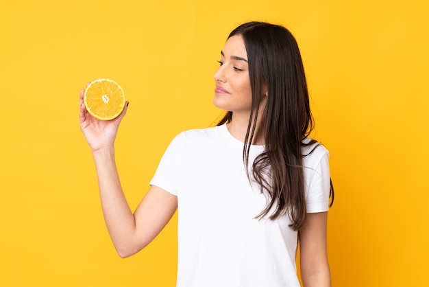 Mujer caucásica joven que sostiene una naranja sobre la pared amarilla aislada