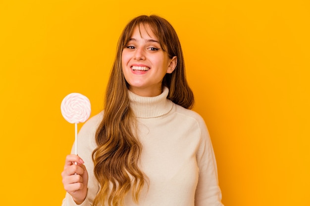 Foto mujer caucásica joven que sostiene una piruleta en amarillo feliz, sonriente y alegre.