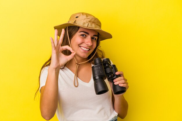 Mujer caucásica joven que sostiene los prismáticos aislados sobre fondo amarillo alegre y confiado que muestra el gesto aceptable.