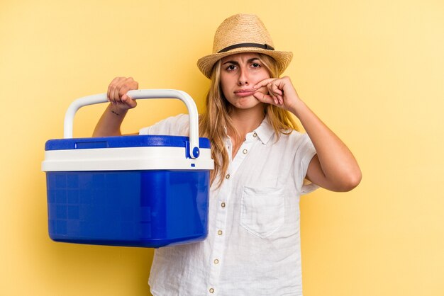 Mujer caucásica joven que sostiene el refrigerador aislado en fondo amarillo con los dedos en los labios que guardan un secreto.