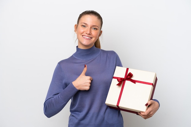 Mujer caucásica joven que sostiene un regalo aislado en el fondo blanco con los pulgares para arriba porque ha sucedido algo bueno