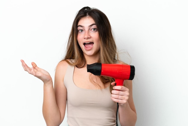 Mujer caucásica joven que sostiene un secador de pelo aislado en el fondo blanco con la expresión facial sorprendida