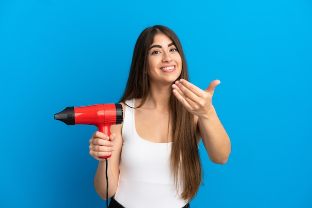 Mujer caucásica joven que sostiene un secador de pelo aislado en la superficie azul que invita a venir con la mano. Feliz de que hayas venido