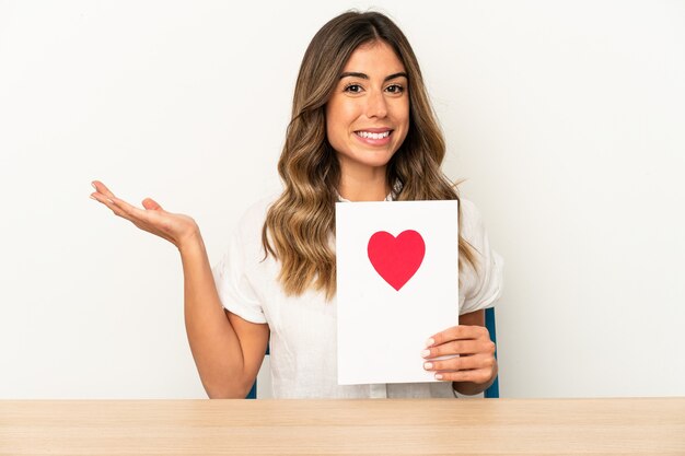 Mujer caucásica joven que sostiene una tarjeta del día de san valentín aislada que muestra un espacio de la copia en una palma y que sostiene otra mano en la cintura.