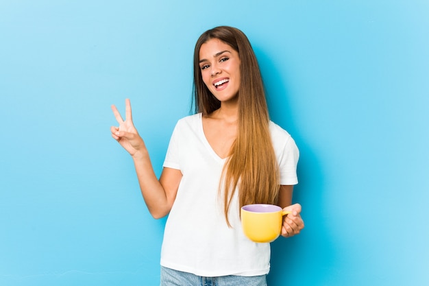 Mujer caucásica joven que sostiene una taza de café alegre y despreocupada que muestra un símbolo de paz con los dedos.