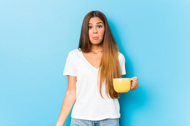 La mujer caucásica joven que sostiene una taza de café encoge de hombros y abre los ojos confundidos.