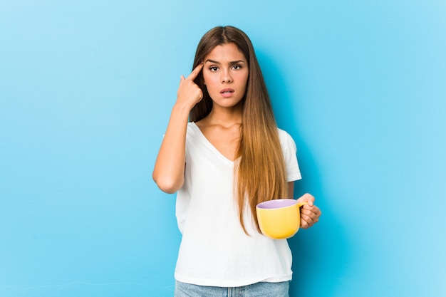 Mujer caucásica joven que sostiene una taza de café que muestra un gesto de decepción con el índice.