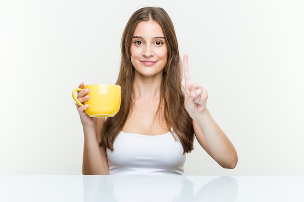 Mujer caucásica joven que sostiene una taza que muestra el número uno con el dedo.