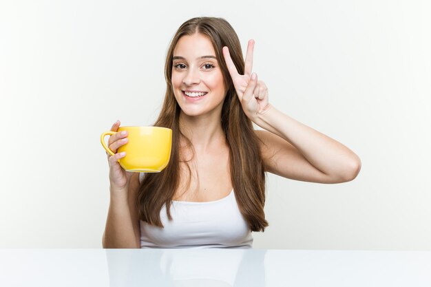 Mujer caucásica joven que sostiene una taza que muestra el signo de la victoria y una amplia sonrisa.