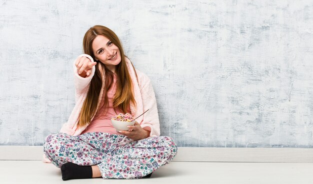 Mujer caucásica joven que sostiene un tazón de cereal sonrisas alegres que señalan al frente.