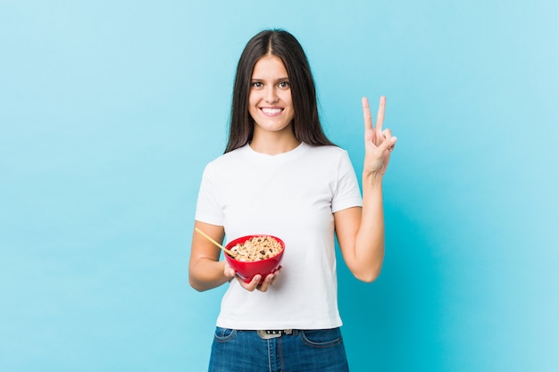 Mujer caucásica joven que sostiene un tazón de cereales que muestra el número dos con los dedos.