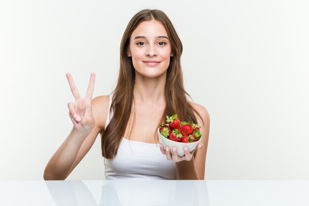 Mujer caucásica joven que sostiene un tazón de fresas que muestra el número dos con los dedos