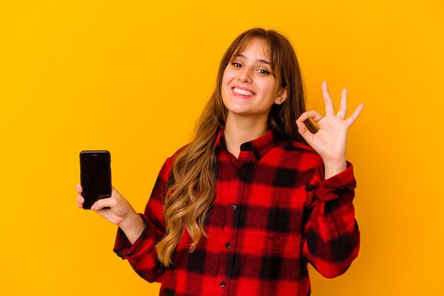 Mujer caucásica joven que sostiene el teléfono aislado en el fondo amarillo alegre y confiado que muestra el gesto aceptable.