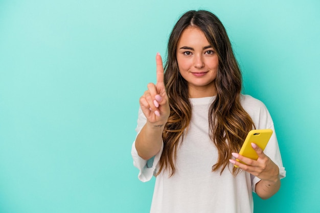 Mujer caucásica joven que sostiene un teléfono móvil aislado sobre fondo azul que muestra el número uno con el dedo.