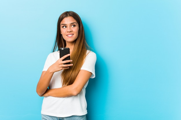 Mujer caucásica joven que sostiene un teléfono que sonríe confiado con los brazos cruzados.