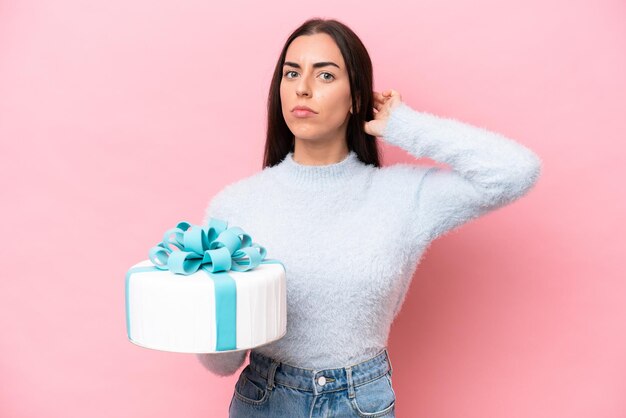 Mujer caucásica joven que sostiene la torta de cumpleaños aislada en el fondo rosado que tiene dudas