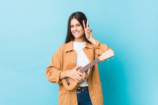 Mujer caucásica joven que sostiene un ukelele que muestra la muestra de la victoria y que sonríe ampliamente.