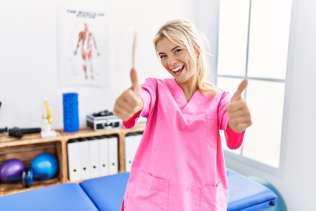 Mujer caucásica joven que trabaja en la clínica de recuperación del dolor aprobando hacer un gesto positivo con los pulgares hacia arriba sonriendo y feliz por el gesto ganador del éxito