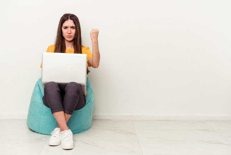 Foto mujer caucásica joven que trabaja con una laptop sentada en un soplo aislado en fondo blanco mostrando el puño a la cámara, expresión facial agresiva.