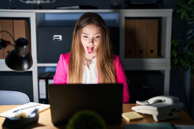 Mujer caucásica joven que trabaja en la oficina por la noche sacando la lengua feliz con expresión divertida. concepto de emoción.