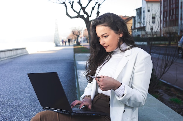 Mujer caucásica joven que usa una tarjeta de crédito roja para pagar en línea con una computadora portátil en la calle