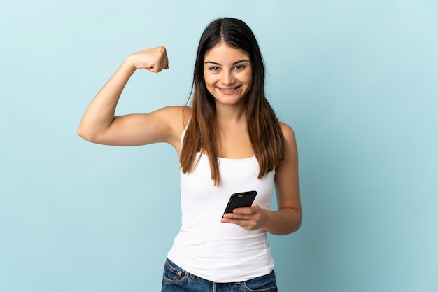 Mujer caucásica joven que usa el teléfono móvil aislado en azul que hace gesto fuerte