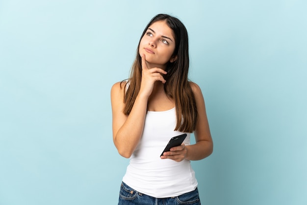 Foto mujer caucásica joven que usa el teléfono móvil aislado en azul que tiene dudas mientras mira hacia arriba
