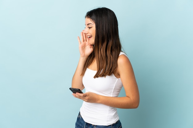 Mujer caucásica joven que usa el teléfono móvil aislado en gritos azules con la boca abierta de par en par al lado