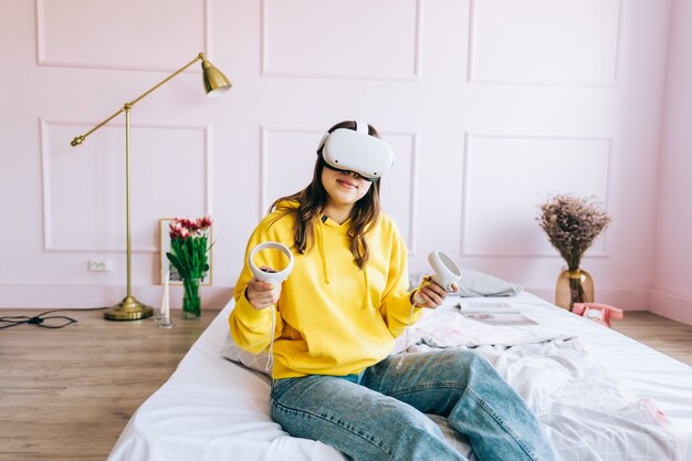 Mujer caucásica joven sentada en la cama y usando auriculares VR, sosteniendo controladores y mirando en realidad virtual.