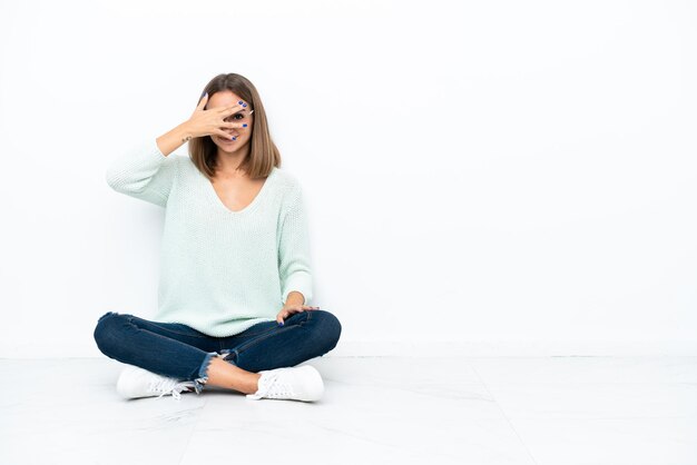 Mujer caucásica joven sentada en el suelo aislada de fondo blanco cubriendo los ojos con las manos y sonriendo