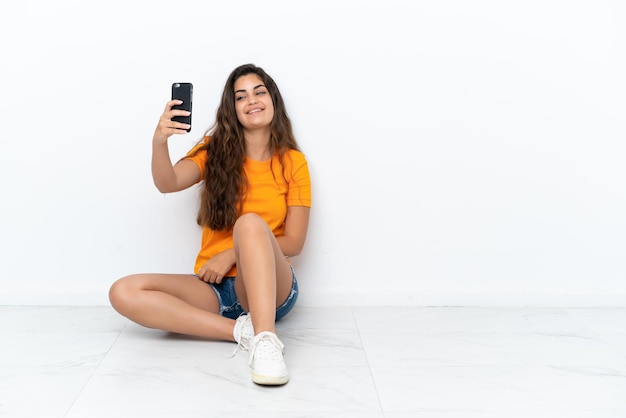 Mujer caucásica joven sentada en el suelo aislada de fondo blanco haciendo un selfie