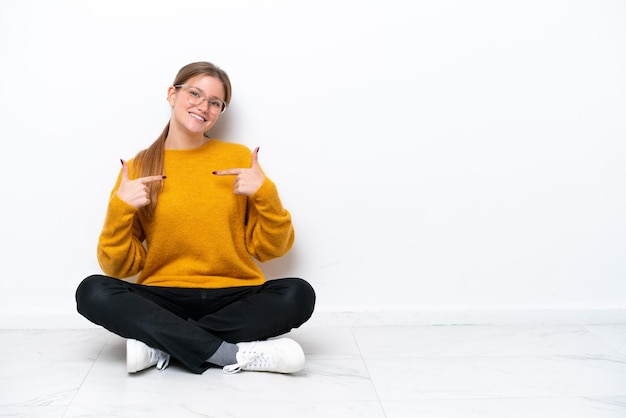 Mujer caucásica joven sentada en el suelo aislada de fondo blanco orgullosa y satisfecha