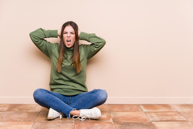 Mujer caucásica joven sentada en el suelo aislado cubriendo las orejas con las manos tratando de no escuchar un sonido demasiado fuerte.