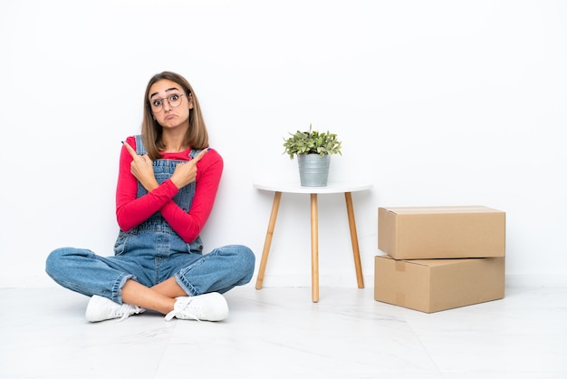 Mujer caucásica joven sentada en el suelo entre cajas apuntando a los laterales con dudas