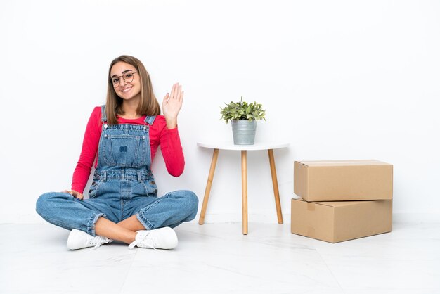 Mujer caucásica joven sentada en el suelo entre cajas saludando con la mano con expresión feliz