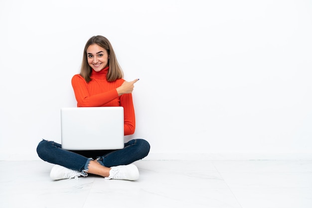 Mujer caucásica joven sentada en el suelo con una computadora portátil apuntando hacia atrás