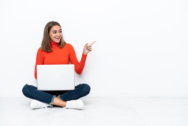 Mujer caucásica joven sentada en el suelo con una computadora portátil apuntando con el dedo hacia un lado y presentando un producto