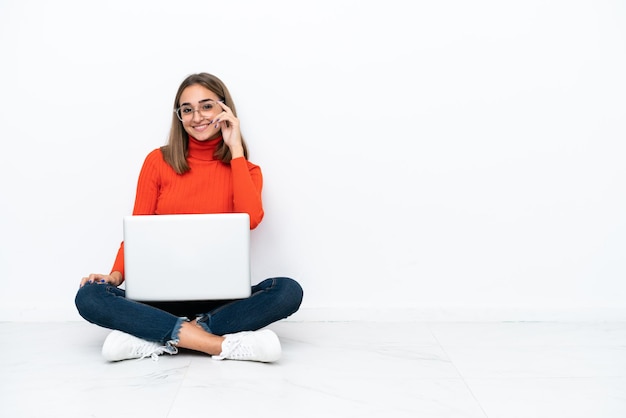 Mujer caucásica joven sentada en el suelo con una computadora portátil con gafas y feliz