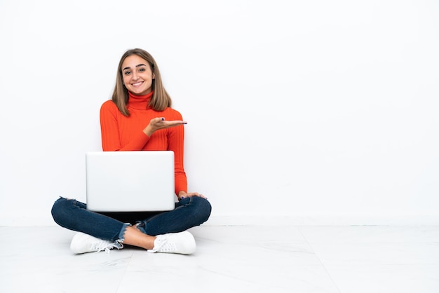 Mujer caucásica joven sentada en el suelo con una computadora portátil que presenta una idea mientras mira sonriendo hacia