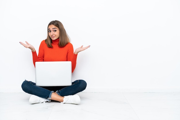 Mujer caucásica joven sentada en el suelo con una computadora portátil que tiene dudas mientras levanta las manos