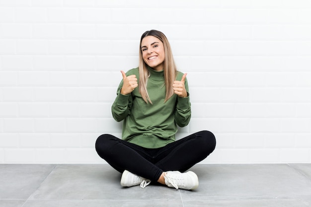 Foto mujer caucásica joven sentada en el suelo levantando ambos pulgares, sonriente y confiado.