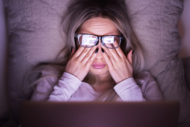 Mujer caucásica joven soñolienta con gafas frotándose los ojos, se siente cansada después de trabajar en una computadora portátil, acostada sobre la almohada, vista frontal