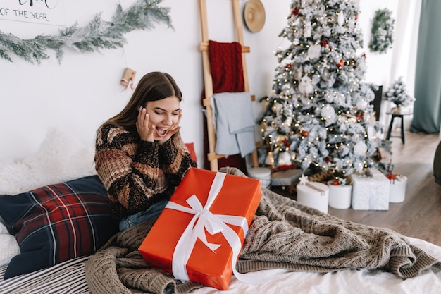 Mujer caucásica joven sorprendida con gran regalo de Navidad mientras está sentado en una cama en casa cerca del árbol de Navidad.