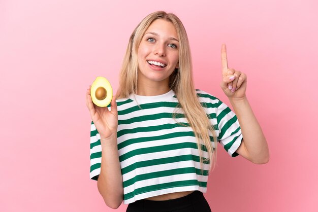 Mujer caucásica joven sosteniendo un aguacate aislado en un fondo rosa señalando una gran idea