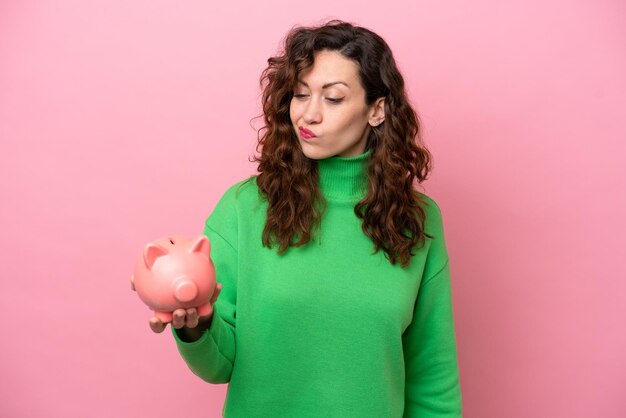 Mujer caucásica joven sosteniendo alcancía aislado sobre fondo de color rosa con expresión triste