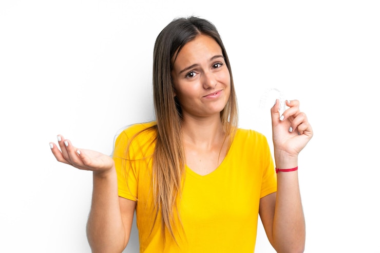 Foto mujer caucásica joven sosteniendo aparatos ortopédicos invisibles aislados en fondo blanco haciendo gestos de duda mientras levanta los hombros