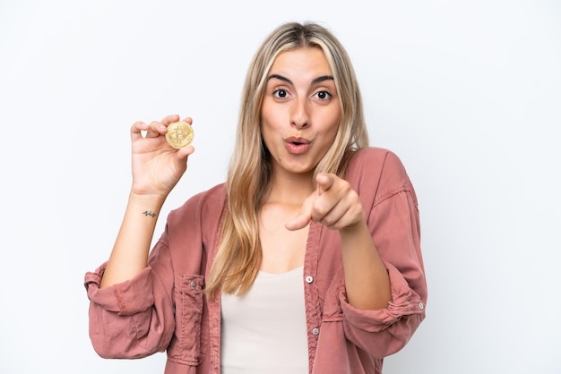 Mujer caucásica joven sosteniendo un Bitcoin aislado sobre fondo blanco sorprendido y apuntando al frente