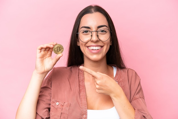 Mujer caucásica joven sosteniendo un Bitcoin aislado sobre fondo rosa y apuntándolo