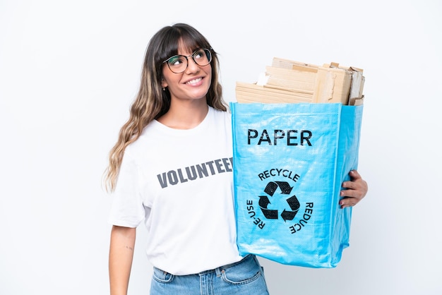 Mujer caucásica joven sosteniendo una bolsa de reciclaje llena de papel para reciclar aislado sobre fondo blanco mirando hacia arriba mientras sonríe